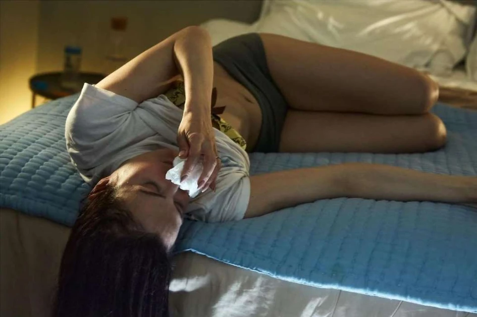 Adult woman lying on a blue bed with white linens, holding a tissue to her face, indoors in a dimly lit room with a side table.