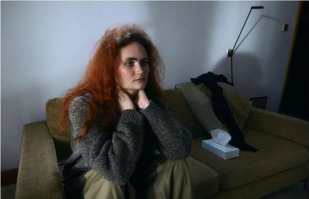 Woman with auburn hair sitting on a sofa indoors wearing a grey sweater and khaki pants. Box of tissues on sofa. Interior shot.