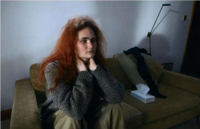 Woman with auburn hair sitting on a sofa indoors wearing a grey sweater and khaki pants. Box of tissues on sofa. Interior shot.