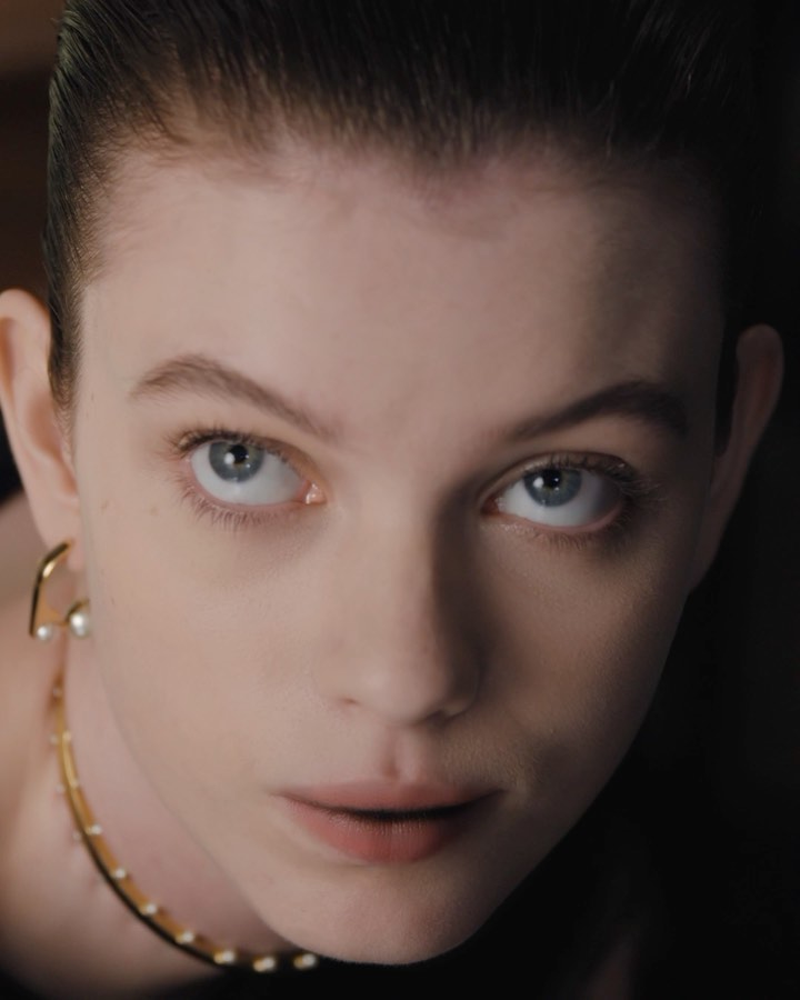 Close-up portrait of a woman with slicked-back dark hair wearing gold earrings and a delicate gold necklace against a dark background.
