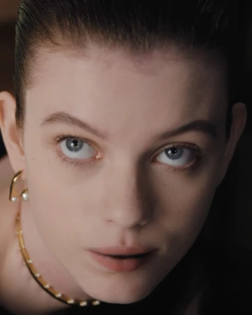Close-up portrait of a woman with slicked-back dark hair wearing gold earrings and a delicate gold necklace against a dark background.