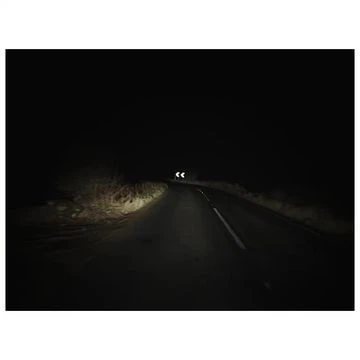 Dark asphalt road with white line markings curving ahead under bright vehicle headlights at night in rural environment.