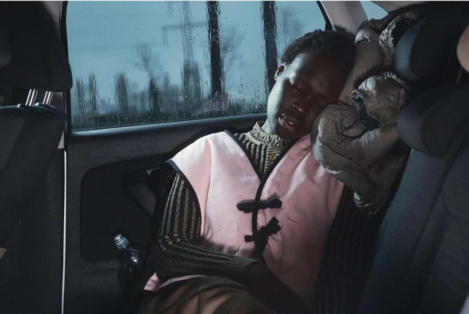 Young adult woman sleeps in the back seat of a car, wearing a pink vest and striped shirt, captured in soft, diffused natural light.