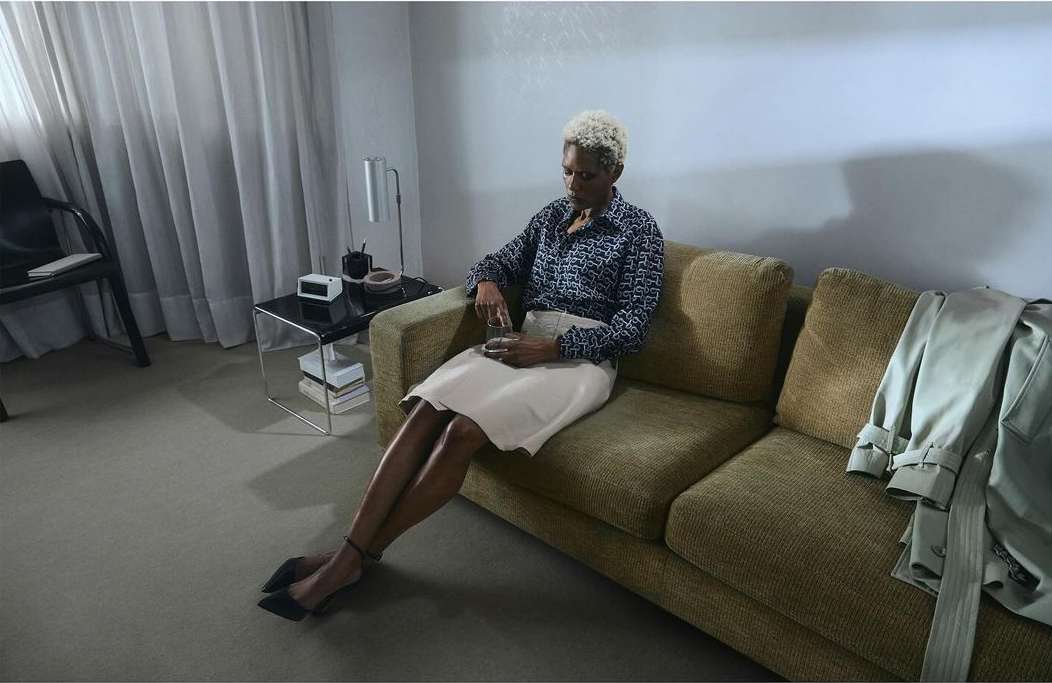 Stylish adult woman sits on an olive-green couch in a modern room with a side table, lamp, coat, and floor-to-ceiling curtains.