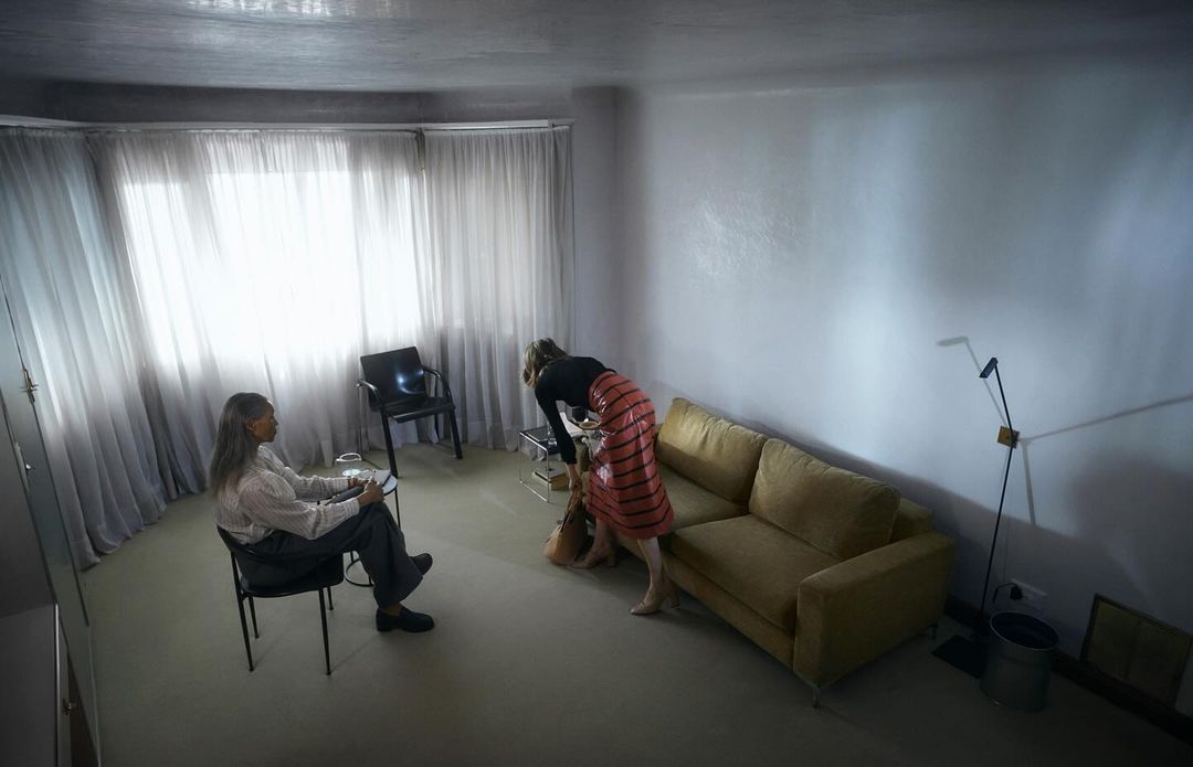 Indoor scene featuring two women in a living room with neutral tones, one seated and the other reaching into a tan tote bag by a couch.