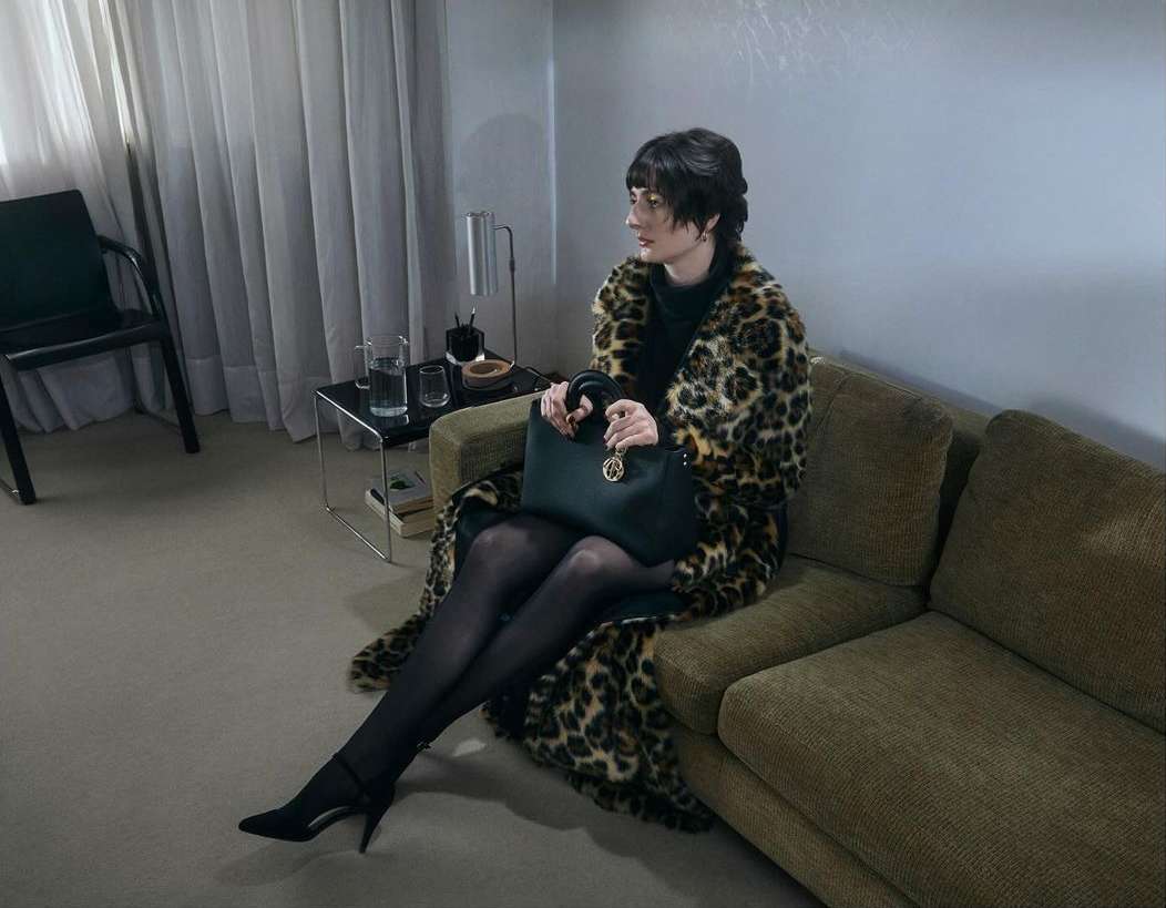 Woman in leopard coat sitting on couch holding black purse in neutral toned indoor setting. She wears black hosiery and heels.