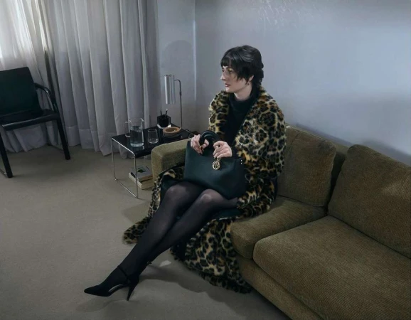 Woman in leopard coat sitting on couch holding black purse in neutral toned indoor setting. She wears black hosiery and heels.