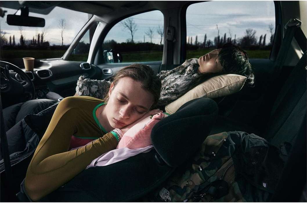 Two people sleeping in a car with pillows and blankets, visible through the windows is a rural landscape with trees and a cloudy sky.