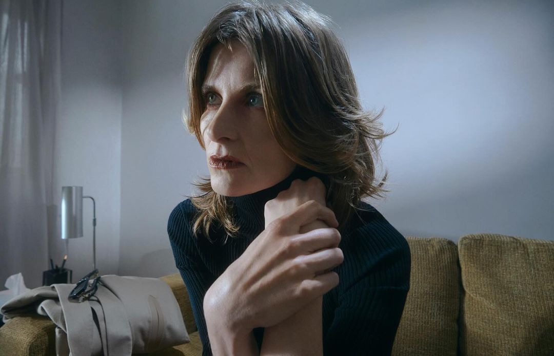 Close-up of a middle-aged woman with short brown hair wearing a black turtleneck sweater, seated on a mustard-colored sofa indoors.