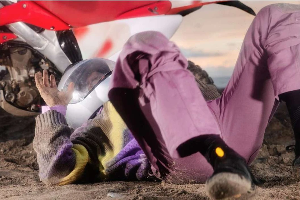 Person in lavender trousers lies beneath a red-and-white motocross bike on sand under a sunset sky in a medium outdoor shot.