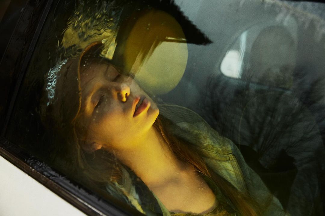 Young woman with closed eyes is seen through a car window with water droplets, reflecting ambient light in an intimate portrait.