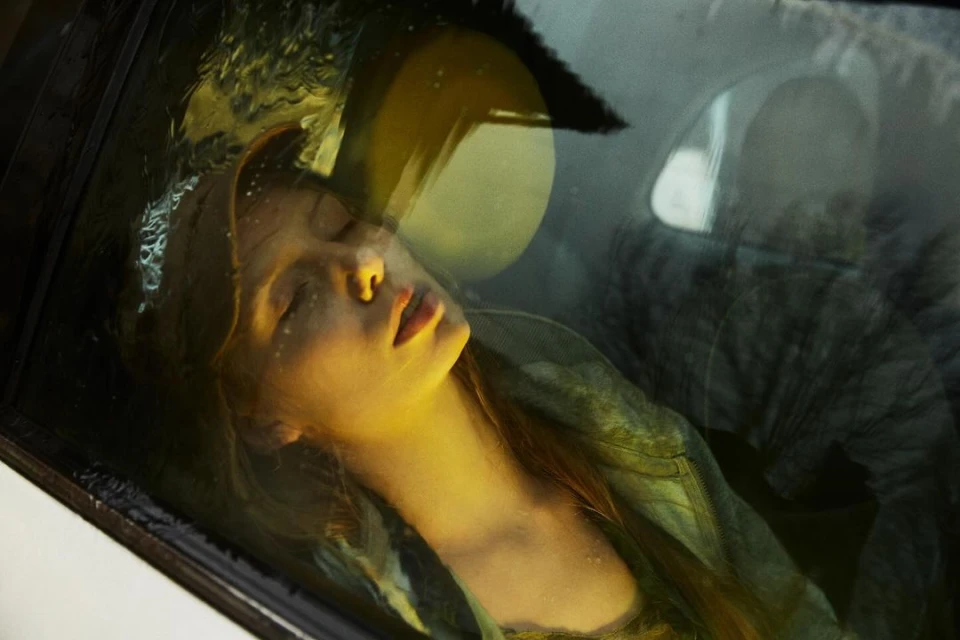 Young woman with closed eyes is seen through a car window with water droplets, reflecting ambient light in an intimate portrait.
