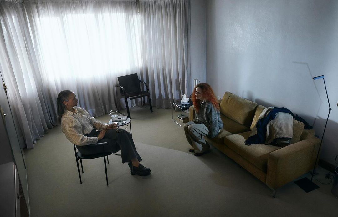 Two women in a counseling session. One woman sits on a couch with a blanket, while the other sits on a chair. Light and airy environment.