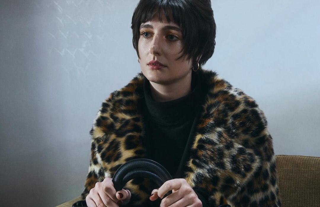 A fair-skinned person with dark hair and a leopard print coat sitting indoors, holding a black purse, looking slightly to the left.