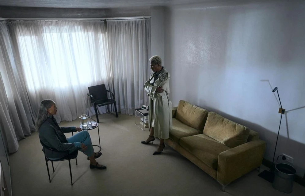 Two adult women in a sparsely decorated interior with soft lighting. One woman sits in a chair while the other stands with crossed arms.