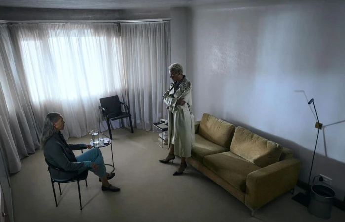 Two women in a sparsely furnished living room, one seated in a chair and the other standing near an olive sofa by sheer curtains.