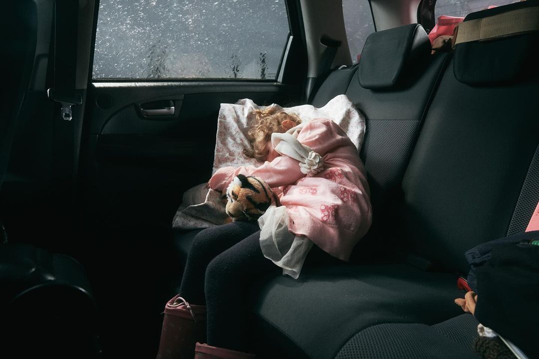 A child is sleeping in the back seat of a car, resting head on a pillow and cuddling a stuffed tiger during a rainy travel day.