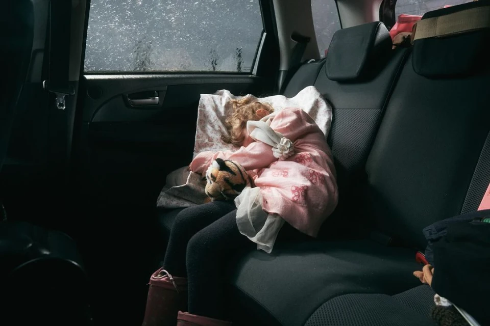 A child is sleeping in the back seat of a car, resting head on a pillow and cuddling a stuffed tiger during a rainy travel day.