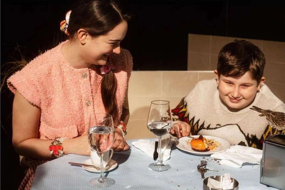 A young girl and boy sit at an indoor restaurant table. The girl wears a pink textured jacket, while the boy has a patterned sweater.