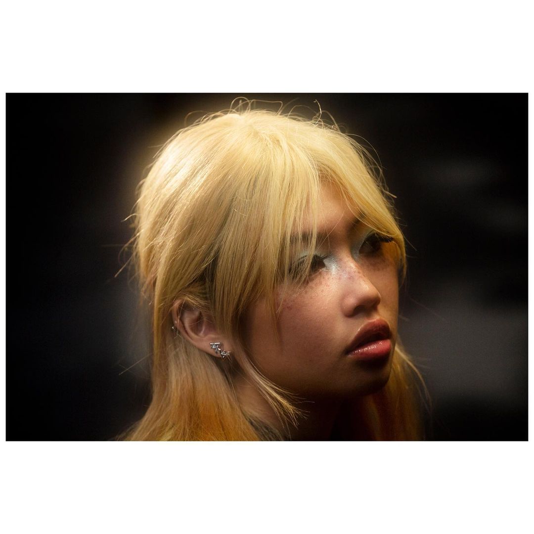 Close-up portrait of a young woman with blonde hair, bangs, and shimmering blue eyeshadow against a dark background.