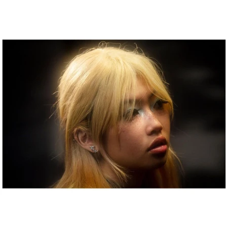 Close-up portrait of a young woman with blonde hair, bangs, and shimmering blue eyeshadow against a dark background.