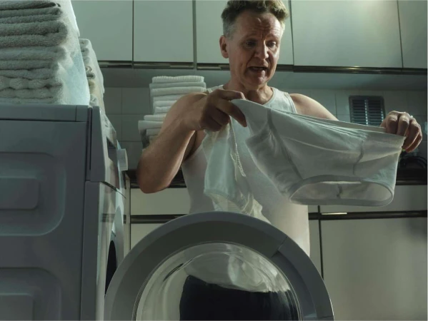 An adult man stands in front of a washing machine in a laundry room, holding a pair of white underwear with a concerned expression.