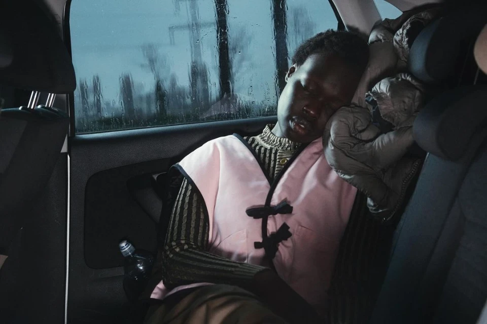 Woman sleeping in the backseat of a car with rain on the window in the background. She wears a pink top, with a gray jacket behind her.
