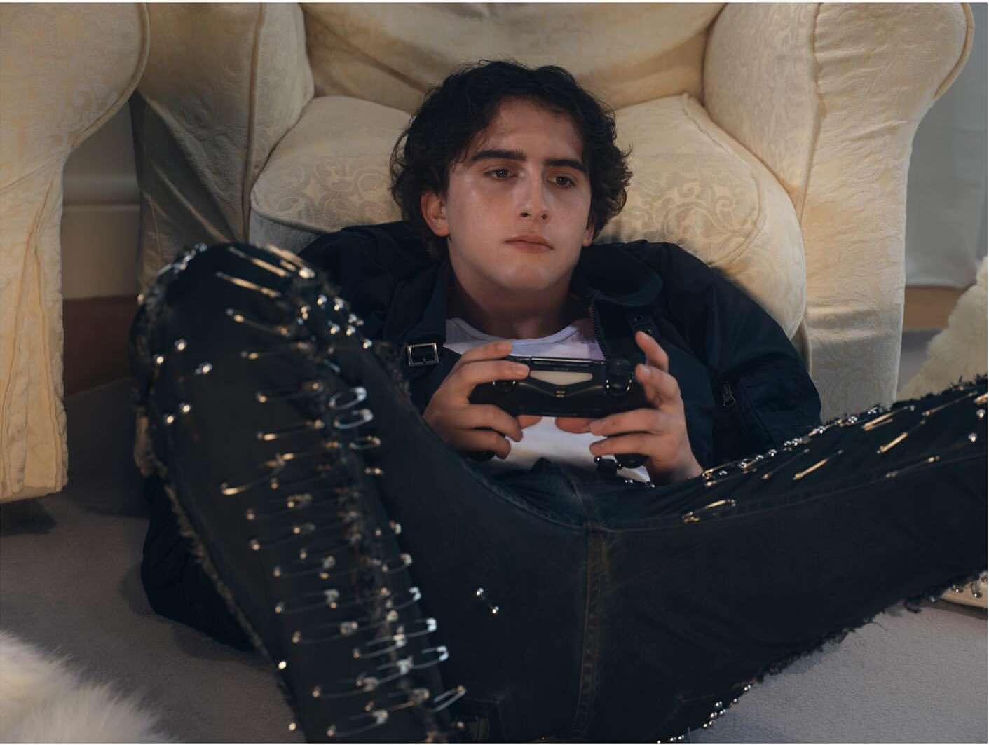 Young man in casual clothing lies back on an armchair and plays a video game, indoors. The armchair is off-white and patterned.