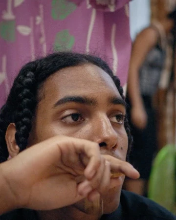 Close-up portrait of a young man with braided hair holding a hand near his mouth, photographed by Alice Schillaci in London.