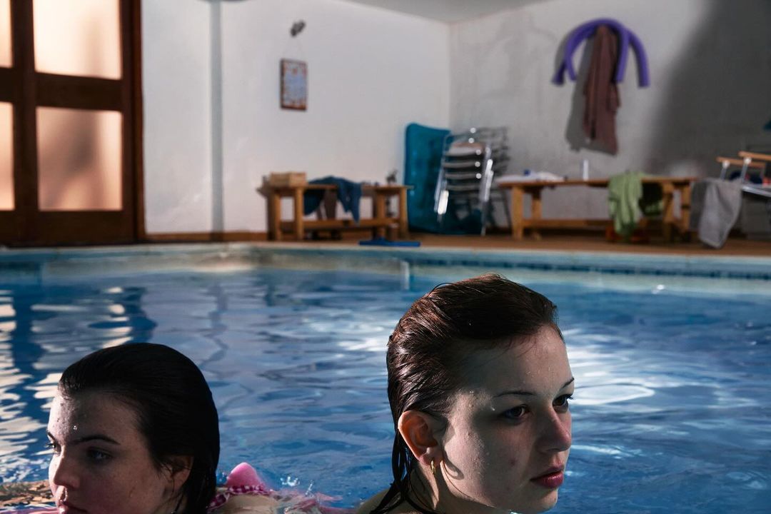 Two young adult females in an indoor swimming pool, with tables and chairs visible in the background of the bright, naturally lit space.