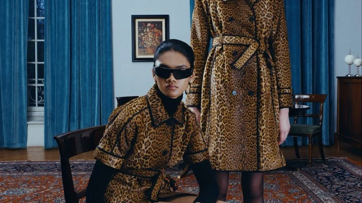 Two women pose indoors wearing matching leopard-print coats with black trim, one seated and one standing in a fashion editorial.