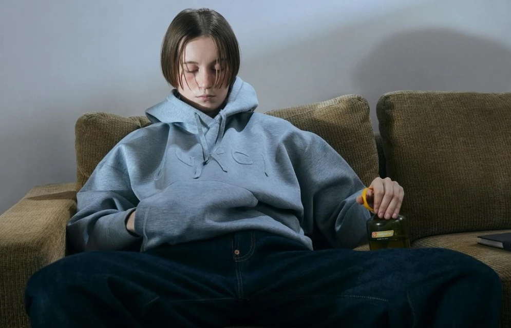 Young woman in a grey hoodie and jeans sits on a couch holding a bottle in an indoor setting with soft lighting.