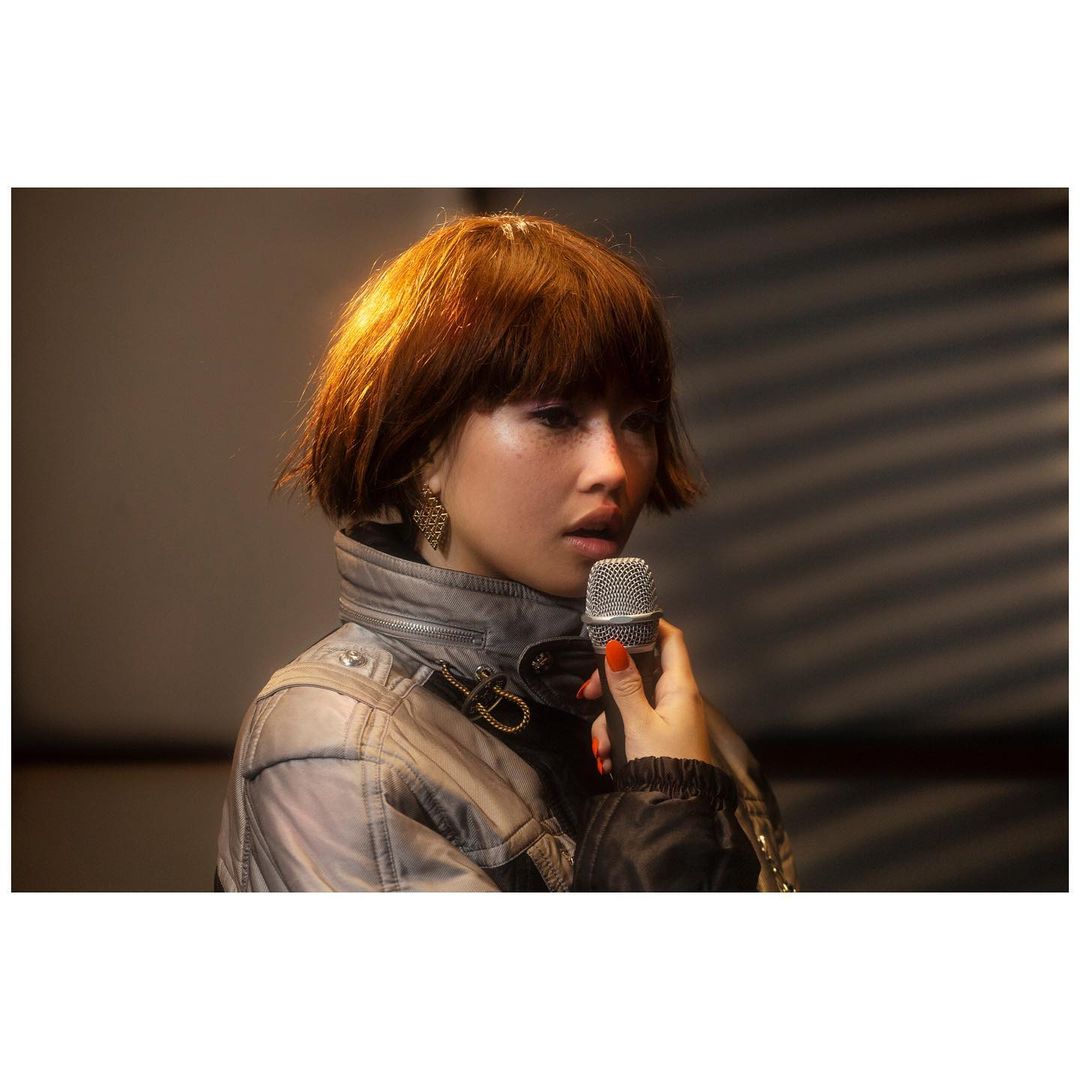 Woman with auburn bob haircut, gray jacket, and orange nail polish holds a microphone in a warm-toned indoor setting.