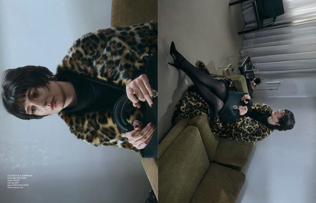A woman in a leopard print coat reclines on a sofa with a handbag, presenting high-fashion style in an indoor setting with soft lighting.