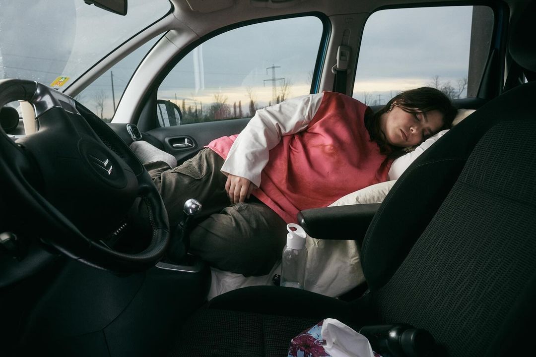 Adult female sleeping inside the front seat of a parked car with a pillow and blanket, visible car interior, and overcast sky.
