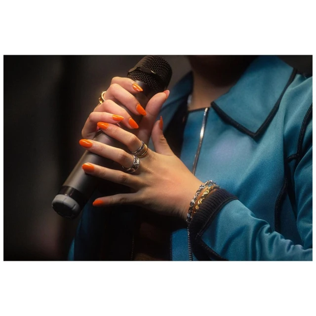 Close up of a woman holding a microphone with orange nails, rings, and bracelets, wearing an aqua blue jacket in a dark environment.