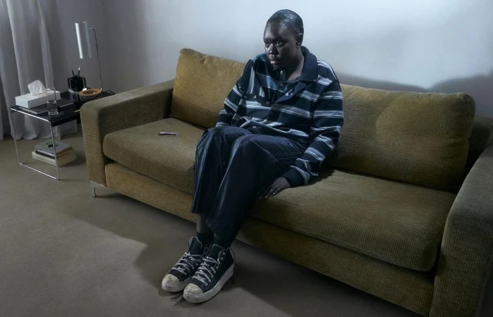 A young adult sits pensively on a couch indoors. The room is sparsely decorated with a modern lamp and side table.