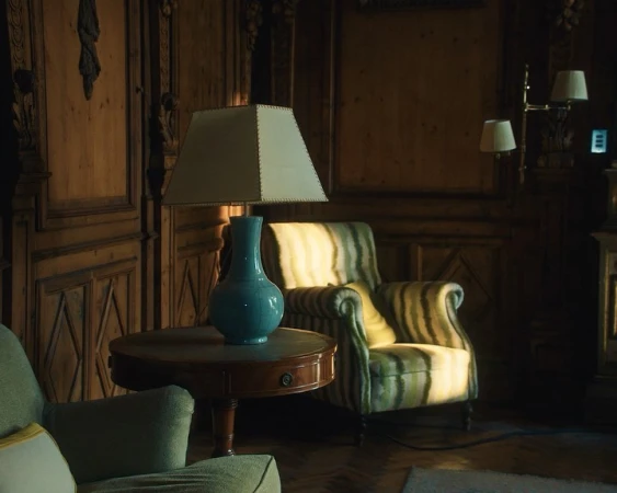 A blue ceramic table lamp on a round wooden table in a wood-paneled room with an armchair in the background.