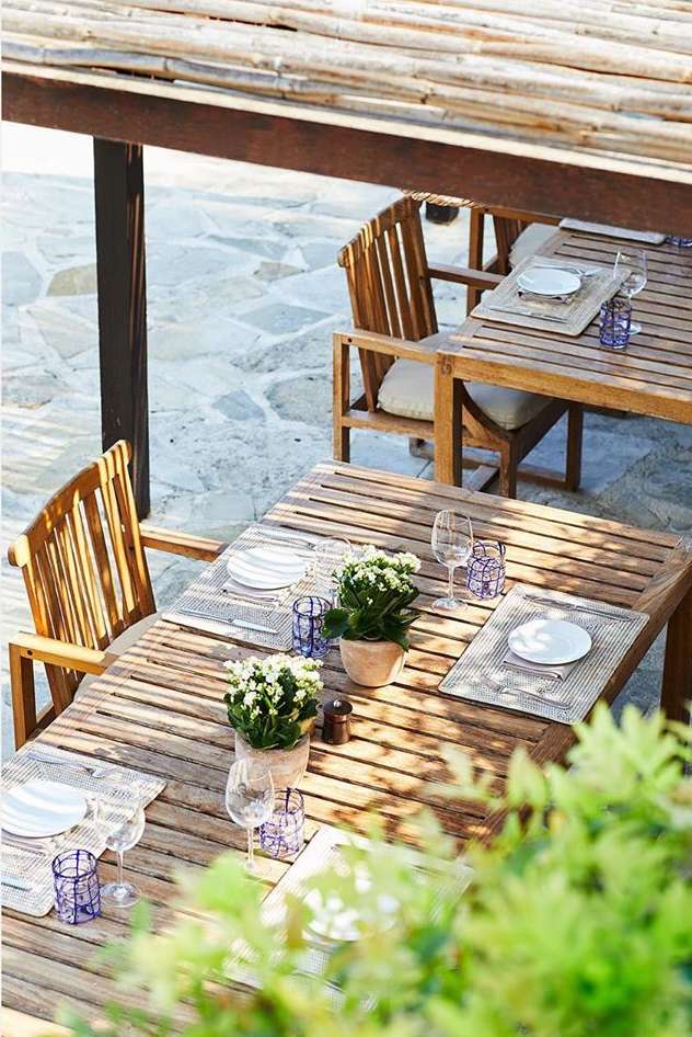 Outdoor dining setting: wooden tables and chairs under a shaded pergola, set with plates, glassware, and potted white flowers.