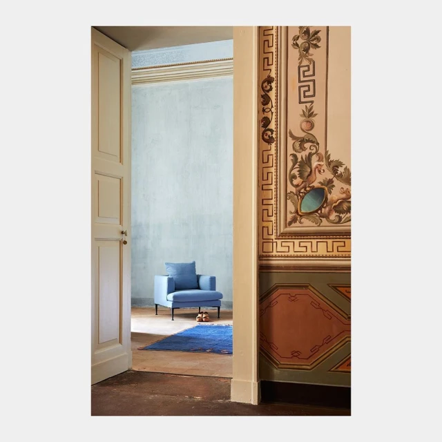 Interior view through doorway of a light blue armchair with black legs, blue rug, and vintage wall art in pastel colors.