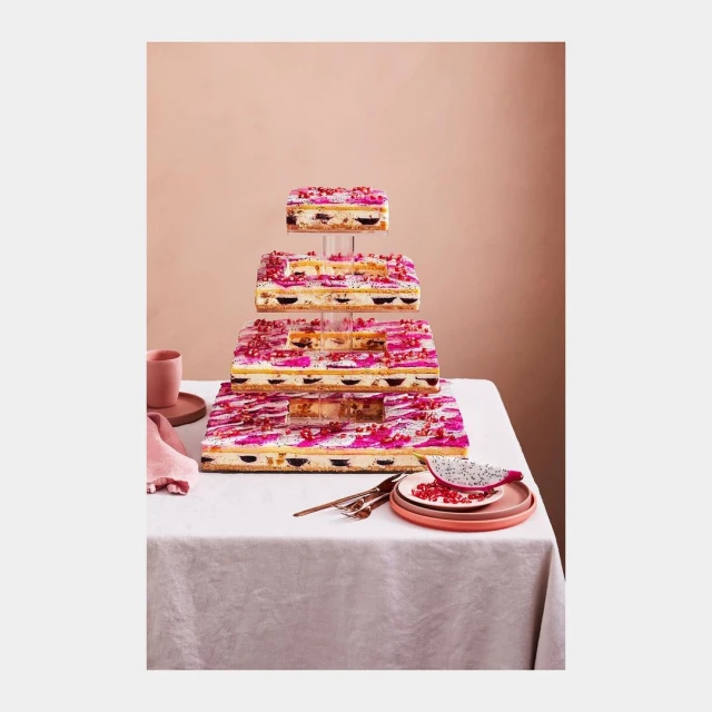 Four-tiered cake with fruit and white frosting on a table with pink accents, against a pastel peach background in a food photography style.