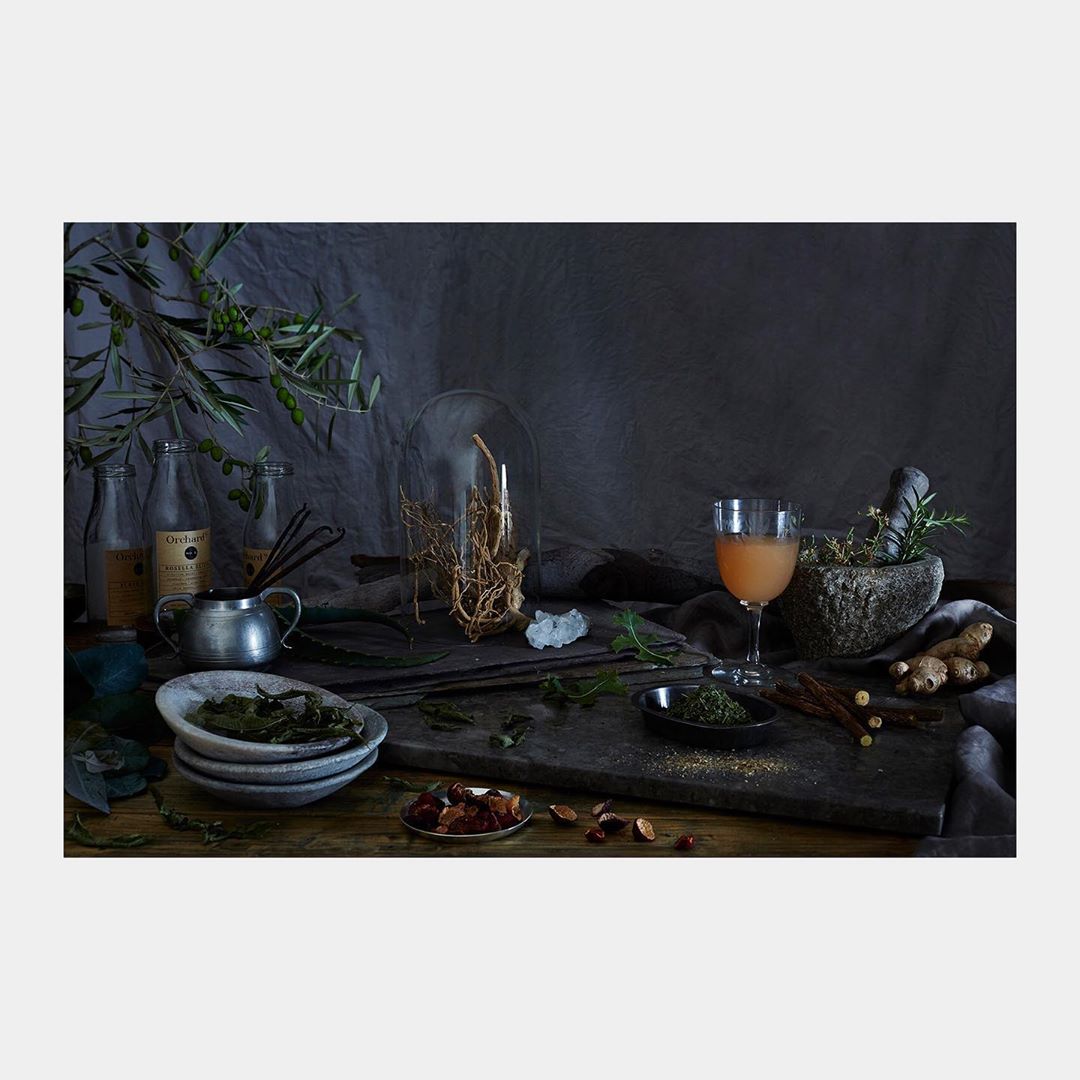 Still life composition with glass bottles, beverage in a glass, olives, herbs, spices, and food ingredients on a dark, rustic wood surface.