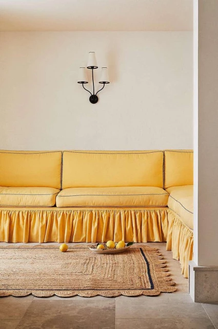 Interior view of a yellow couch with lemons in a woven bowl on a jute rug. Wall sconce on white wall behind the couch.