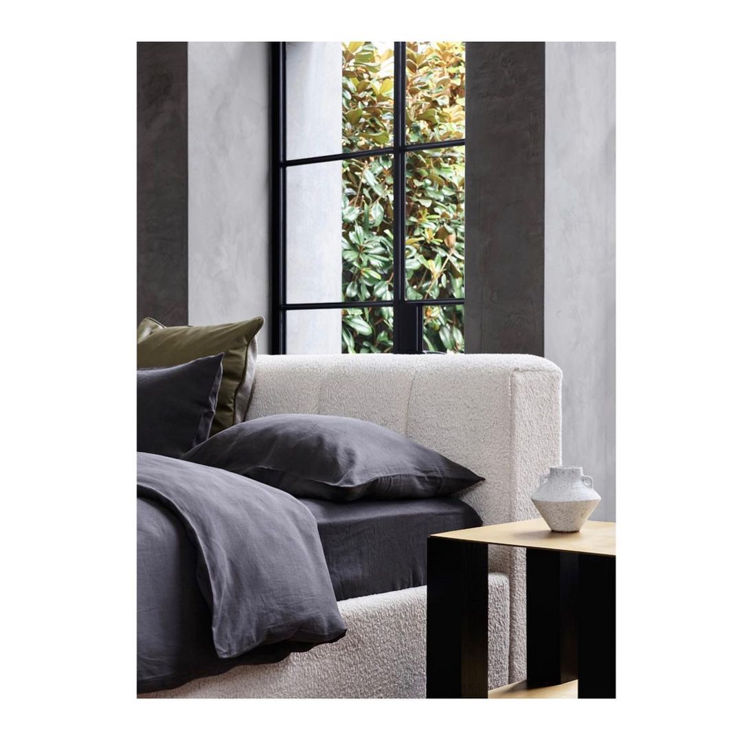Bedroom interior with bed dressed in gray linen, pillows, and a white vase on a wood and black side table near a window.