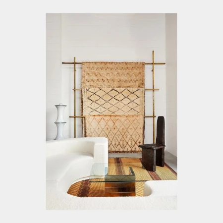 Interior design with white couch, clear glass table, geometric wool rug hanging on gold stand, and dark wood chair against white wall.