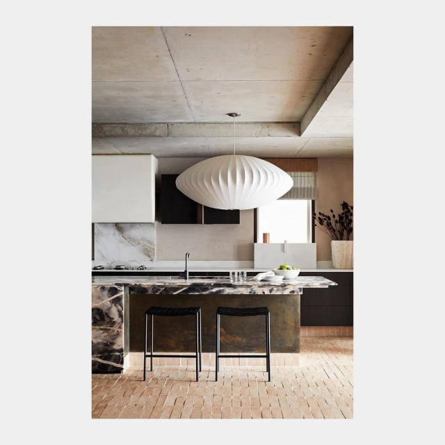 Modern kitchen interior featuring an island with marble countertop, bar stools, unique white pendant light, and light-colored brick flooring.