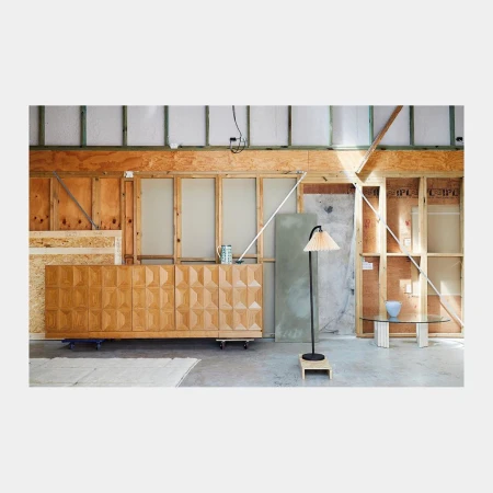 Interior with wooden cabinet and lamp in a construction-like space, featuring glass table with bowl and plywood walls, creating a minimalist aesthetic.