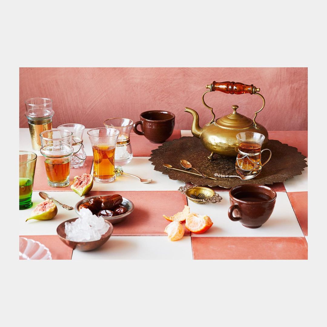 Still life with golden teapot, glasses of tea, dates, figs, and orange slices on a pink and white tiled surface.
