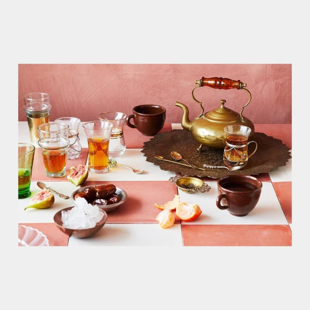 Still life with golden teapot, glasses of tea, dates, figs, and orange slices on a pink and white tiled surface.