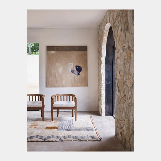 Interior with two wooden chairs, an abstract painting, stone wall detail, and a patterned area rug in neutral tones and natural light.